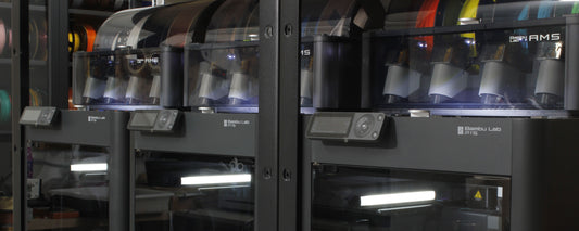 Vista de impresoras 3D BambuLab P1S y sistema AMS en un entorno de taller, mostrando filamentos de colores y varios objetos impresos, destacando la avanzada tecnología de impresión 3D disponible en Jutlo Studio.