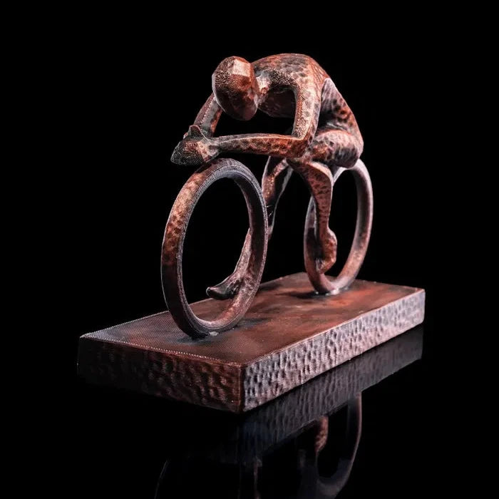 Escultura de un ciclista inclinado hacia adelante en plena carrera, vista frontal, mostrando la intensidad del movimiento en un diseño estilizado de bronce envejecido.